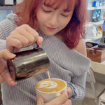 Person pouring milk into a cup of coffee with latte art, in a casual indoor setting.