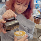 Person pouring milk into a cup of coffee with latte art, in a casual indoor setting.