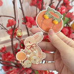 A hand holds two decorative paper-cut style ornaments: a peach-shaped ornament with small yellow pom-poms and a bunny ornament with gold and pearl details, in front of a blurred background of red flowers and branches.