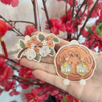 A hand holds two decorative jewelry pieces against a background of red artificial flowers. One piece is a floral sticker or charm with orange blossoms and green leaves, and the other is a pair of dangling earrings with gold-toned frames, clear stones, and white pearl-like drops, displayed on a round illustrated backing card featuring a cartoon rabbit.