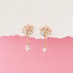A pair of gold-toned dangle earrings with butterfly and teardrop crystal details, displayed against a split cream and pink background.