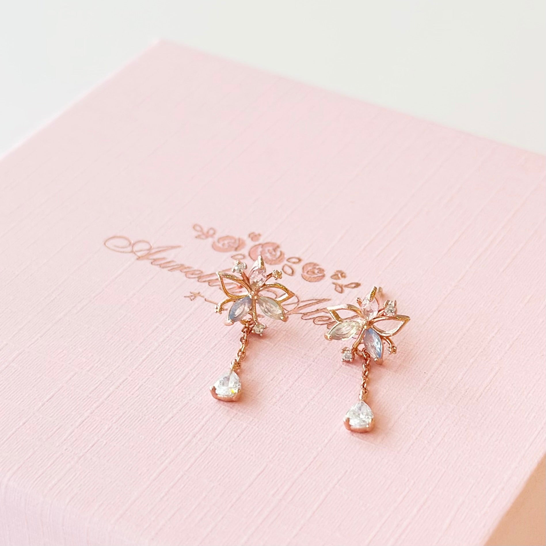A pair of rose gold butterfly dangle earrings with clear rhinestones, resting on a textured light pink gift box with gold foil script.