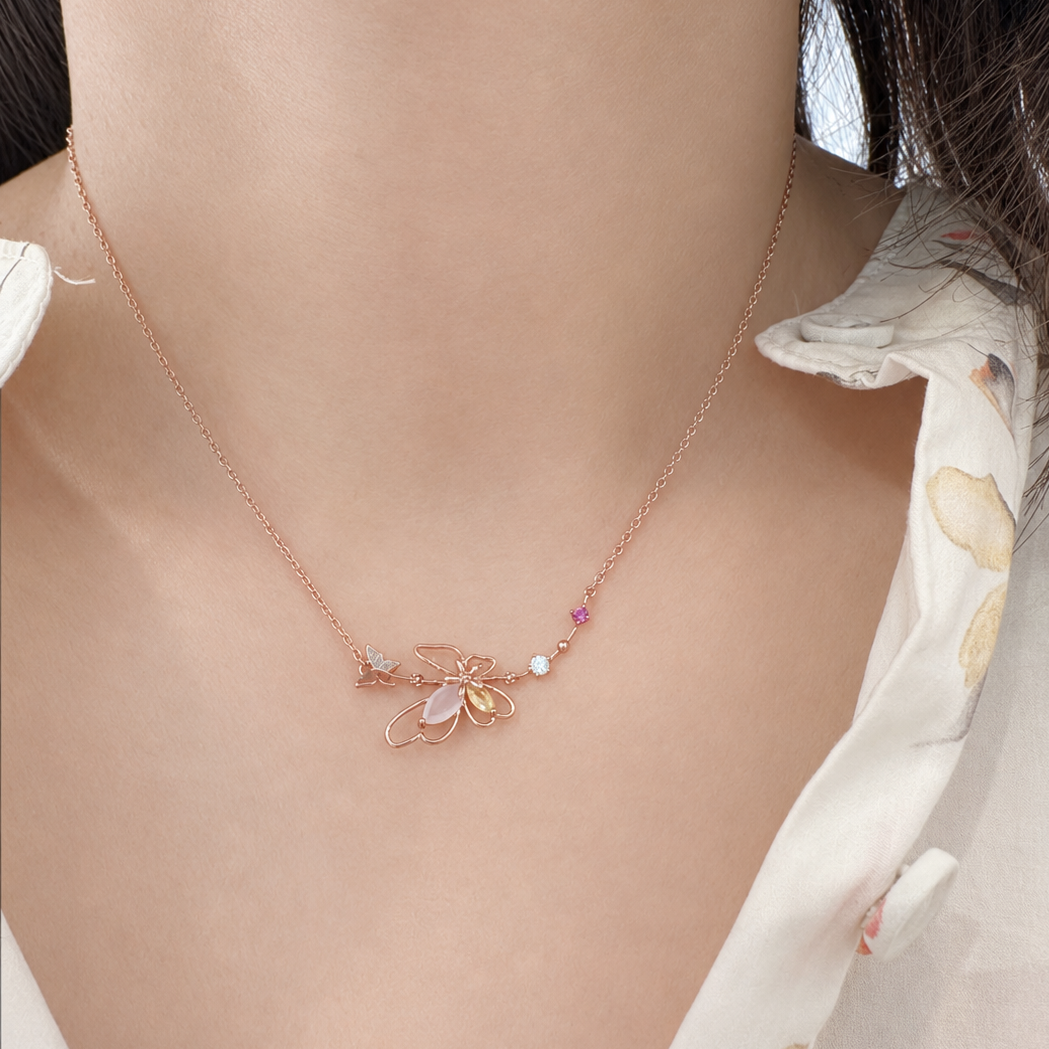A close-up of a model's neck and chest wearing a delicate rose gold necklace with a butterfly pendant, a small white pearl, and a tiny pink gemstone.