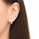 A close-up of a model's ear wearing a slim silver hoop earring set with small clear stones, against a clean white background.