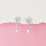 A pair of silver-toned dangle earrings with iridescent light blue and green crystal details, displayed against a split white and pink background.
